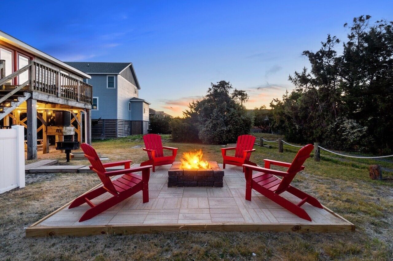 A fire pit with four red Adirondack chairs arranged around it, set on a wooden deck in front of a house with a balcony.