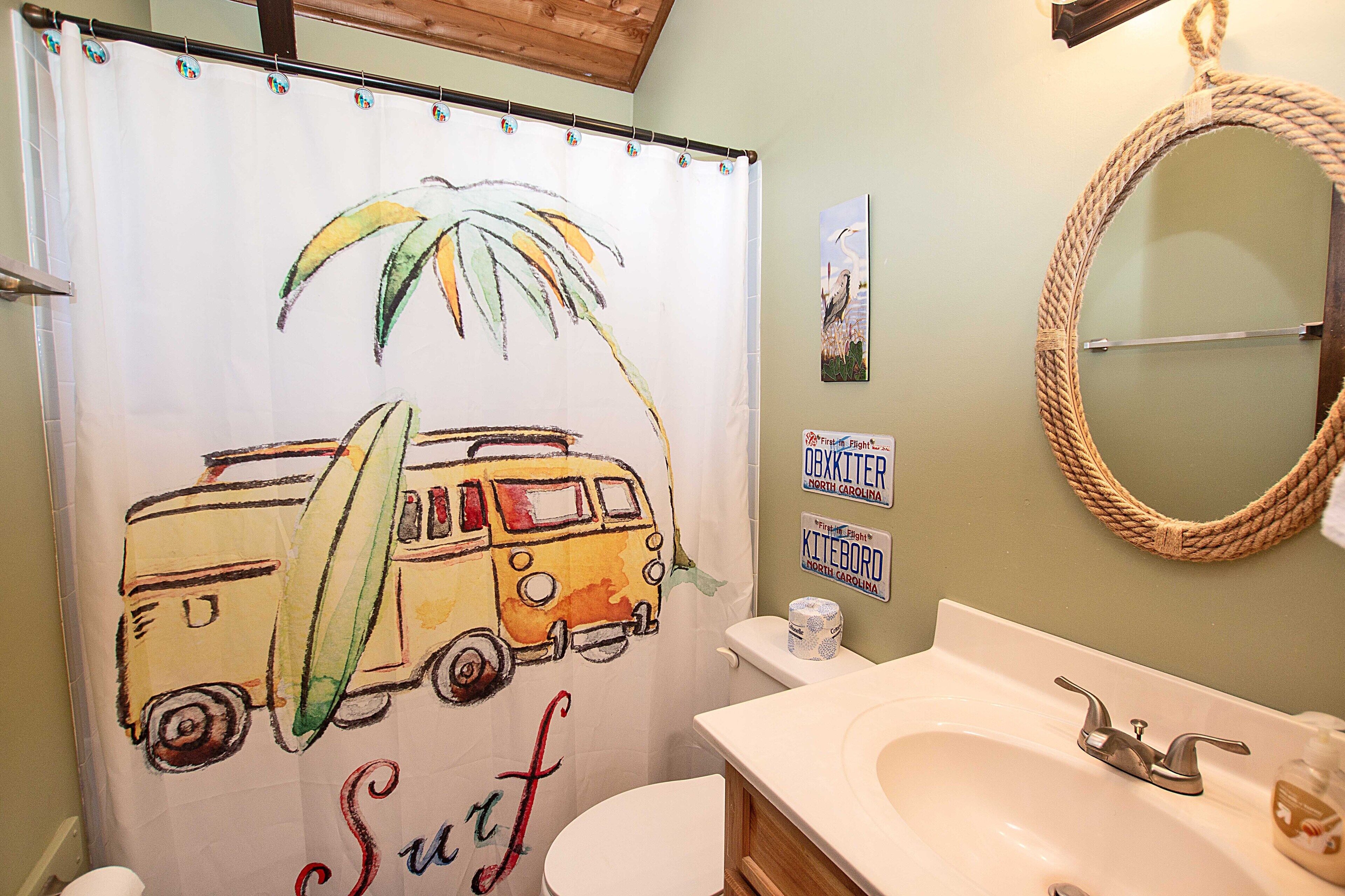 A bathroom with a shower curtain featuring a surfboard and a van, a toilet, a sink with a soap dispenser, and a mirror with a rope frame.