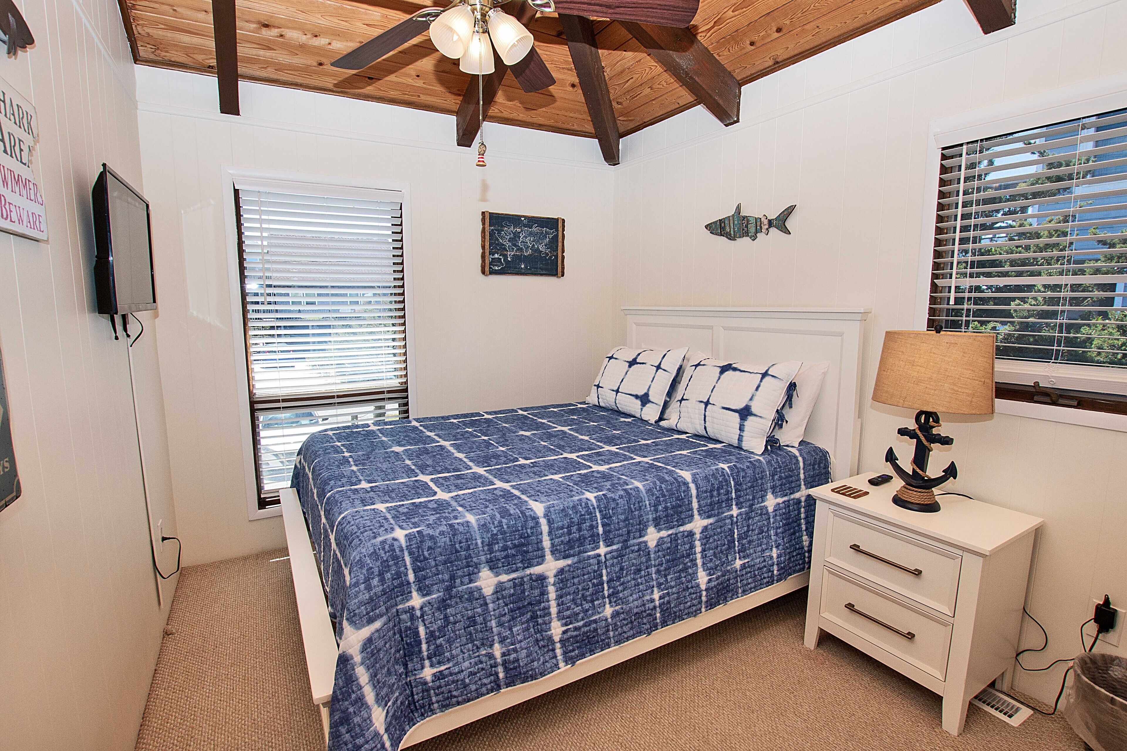 A bedroom with a bed, a nightstand, a wall-mounted TV, a window with blinds, and a ceiling fan.