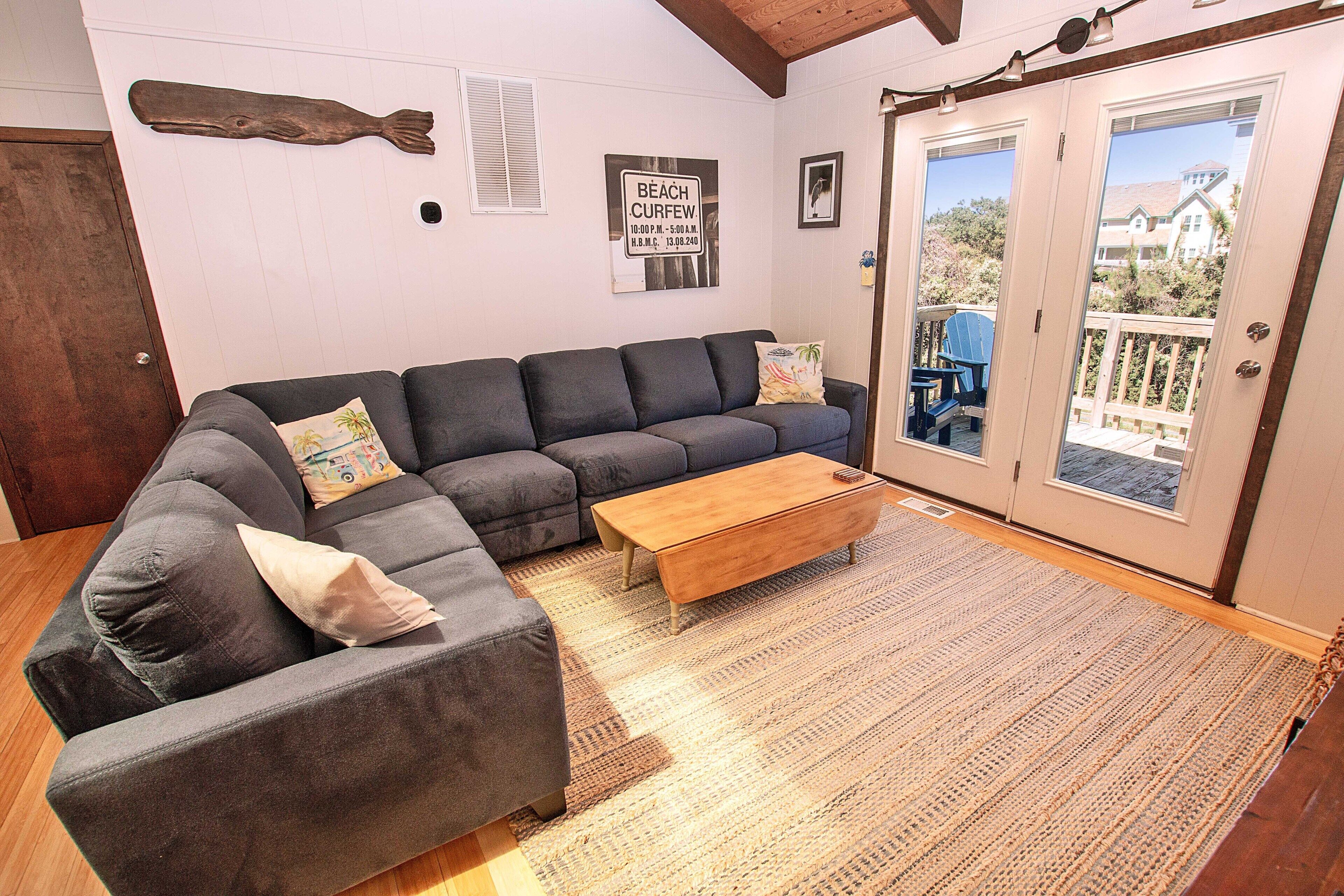 A living room with a grey sectional sofa, a wooden coffee table, and a patterned rug. There is a door leading to a balcony with a view of a house and trees.