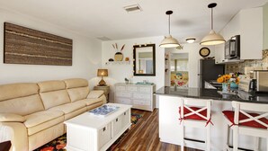 A modern living room with a beige sofa, a white coffee table, and a kitchen area with a bar counter and white bar stools. - Vibrant 1bed/1bath Golf Course Front in the Meadows (Sarasota)