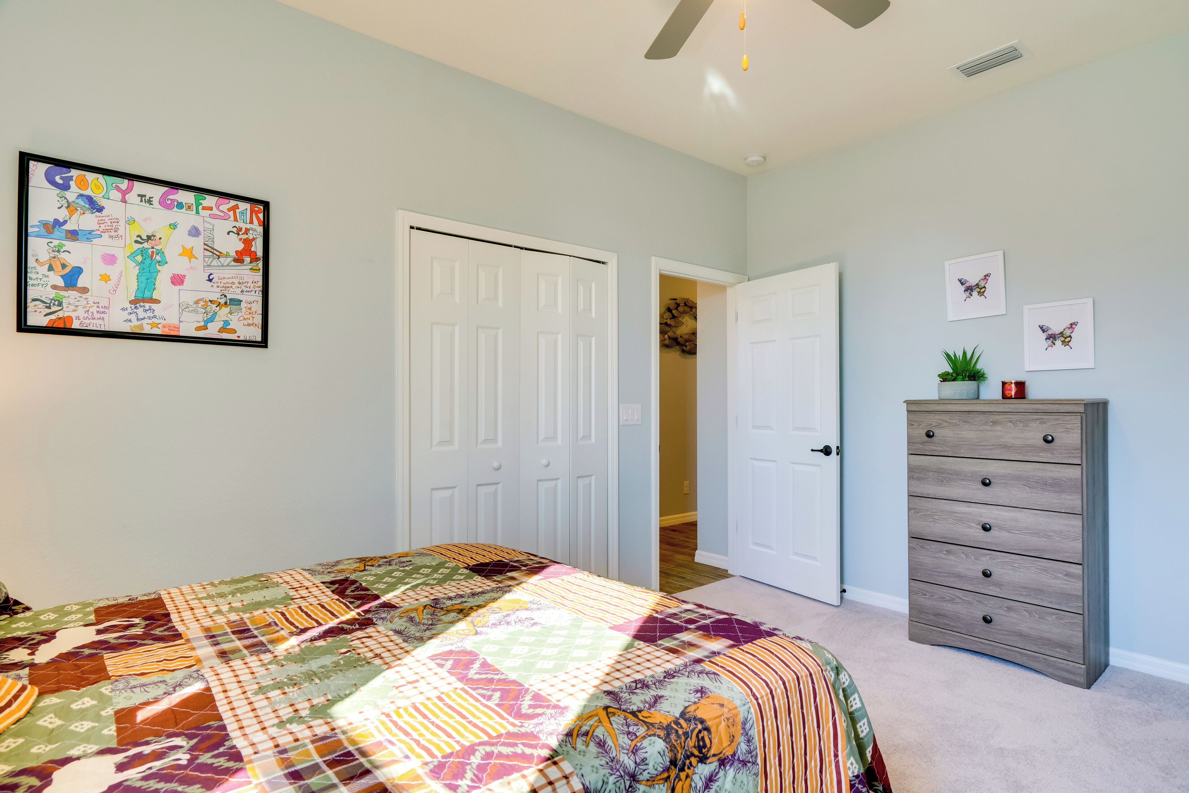 A bedroom with a bed, a dresser, a ceiling fan, and a door leading to another room.