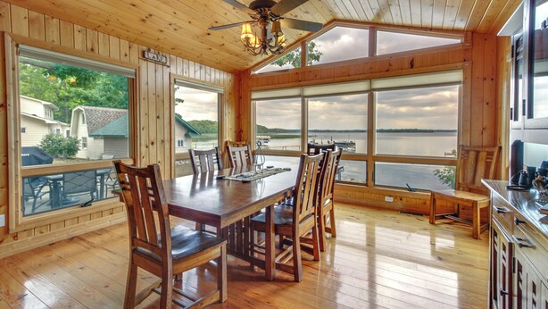 Un comedor de madera con una mesa grande y sillas, un ventilador de techo y ventanas amplias con vistas a un lago.