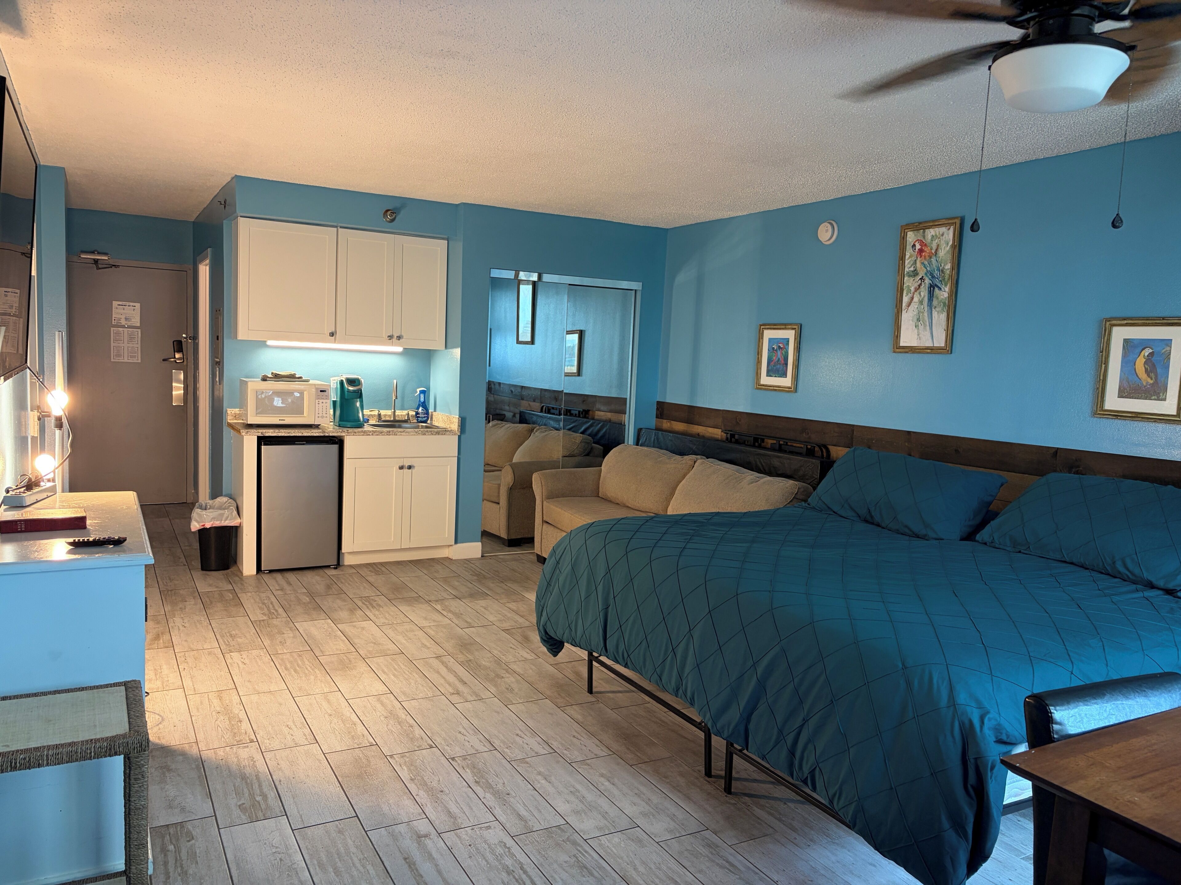 A compact room with a bed, kitchenette, and a ceiling fan.