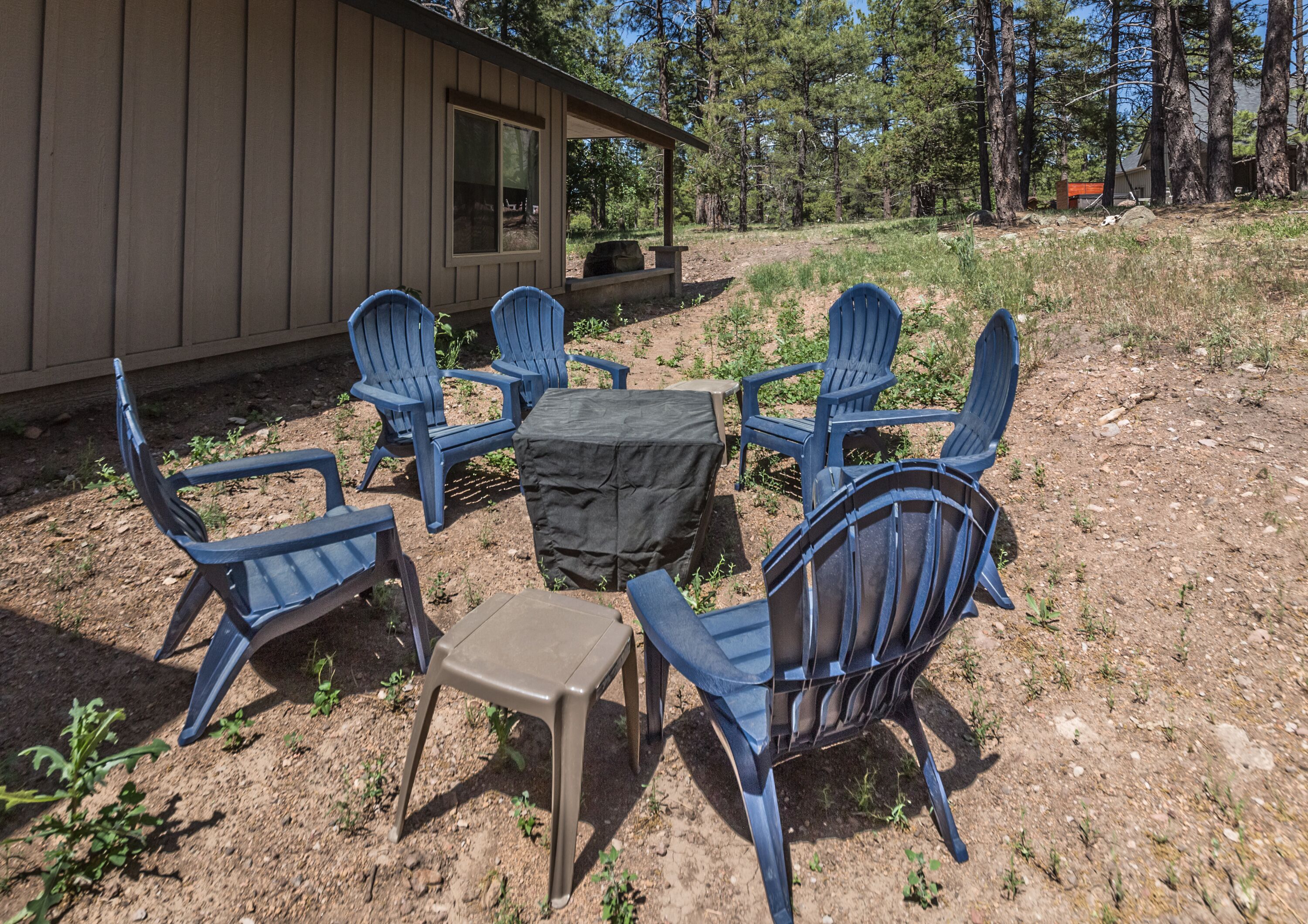 An outdoor seating area with six blue Adirondack chairs and a small table, surrounded by a dirt ground with sparse vegetation.