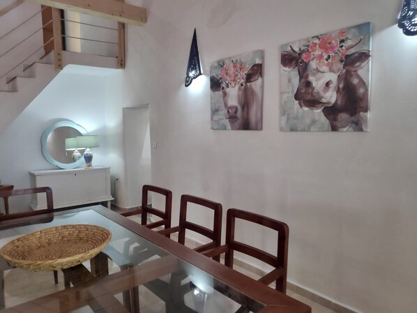 Un comedor con una mesa de vidrio, sillas de madera, una cesta tejida y dos cuadros colgados en la pared de vacas.