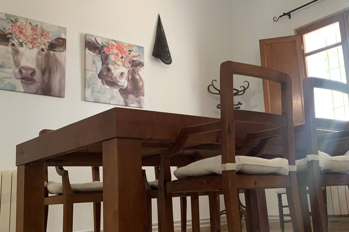 Un comedor con una mesa y sillas de madera, dos cuadros en la pared de vacas, un adorno negro y una ventana con vistas.