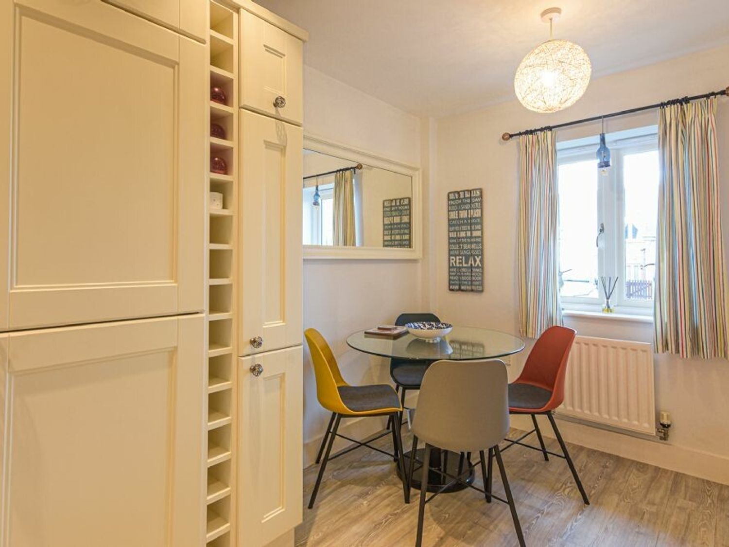 Un coin repas comprenant une table en verre, quatre chaises, un meuble blanc avec des étagères, un miroir et une fenêtre avec des rideaux.