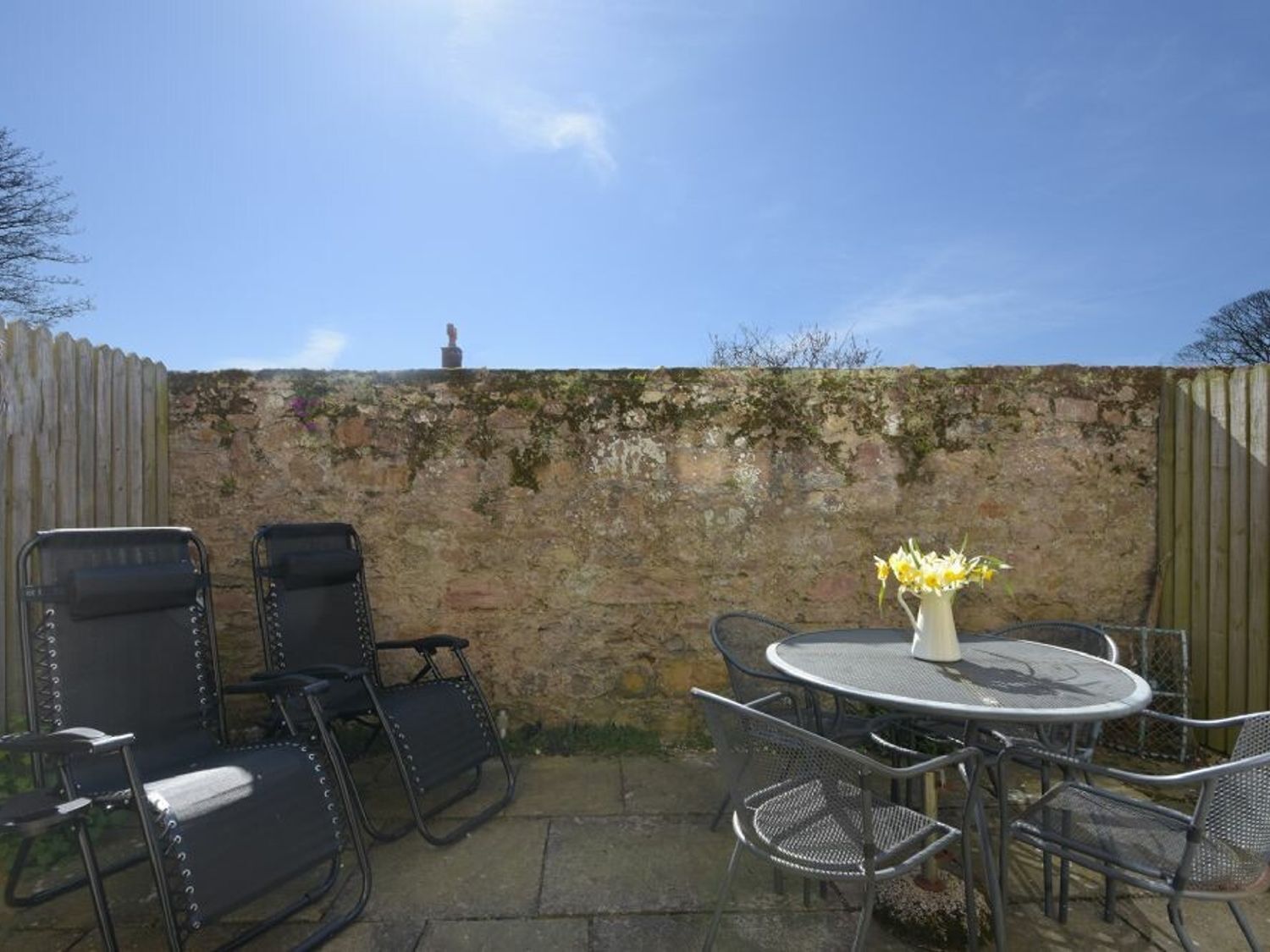 An outdoor patio with metal chairs and a round table, a stone wall, and a small statue.