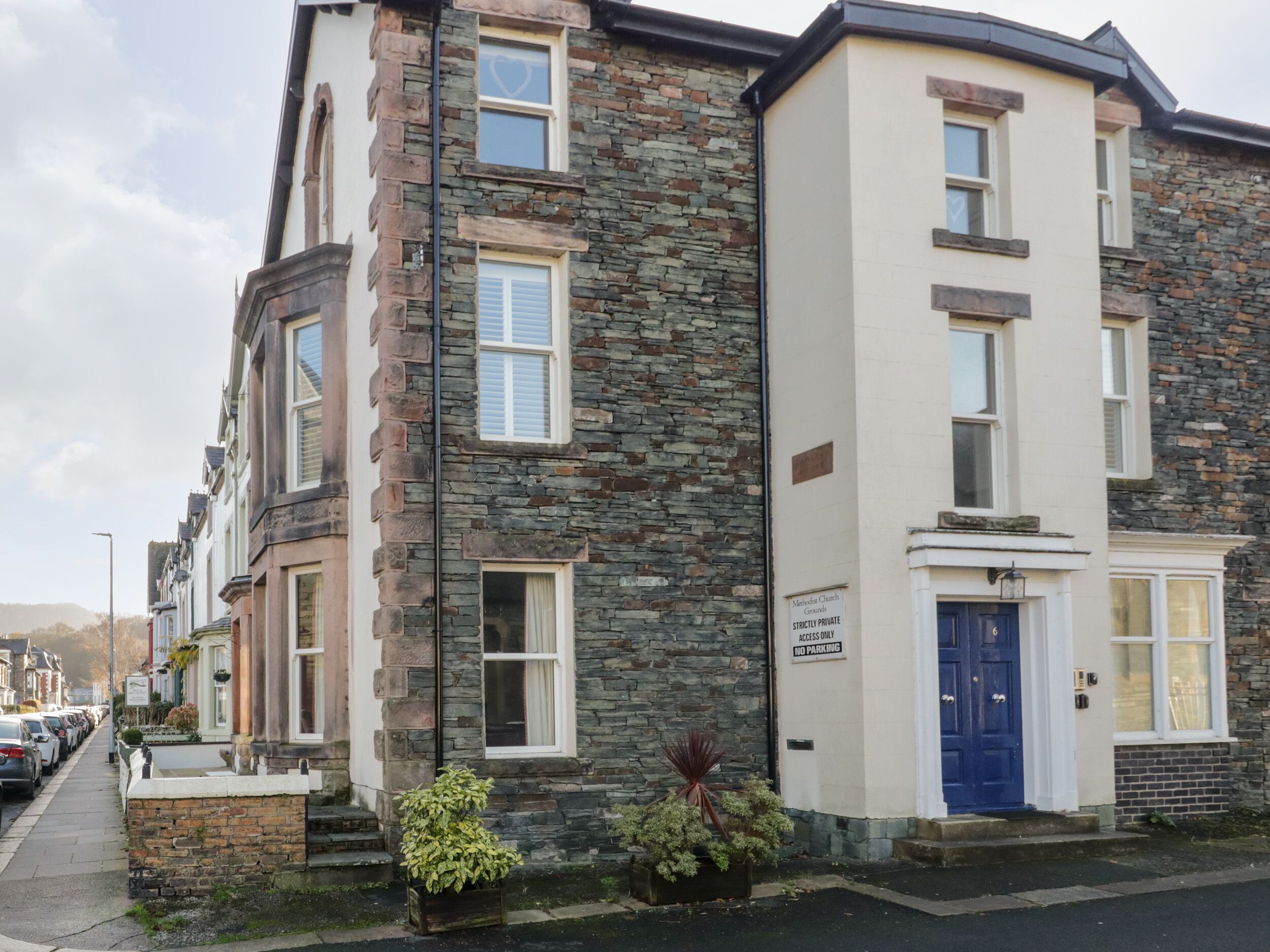 A two-story stone building with a blue door, a sign indicating it is private property, and a row of parked cars along the street.