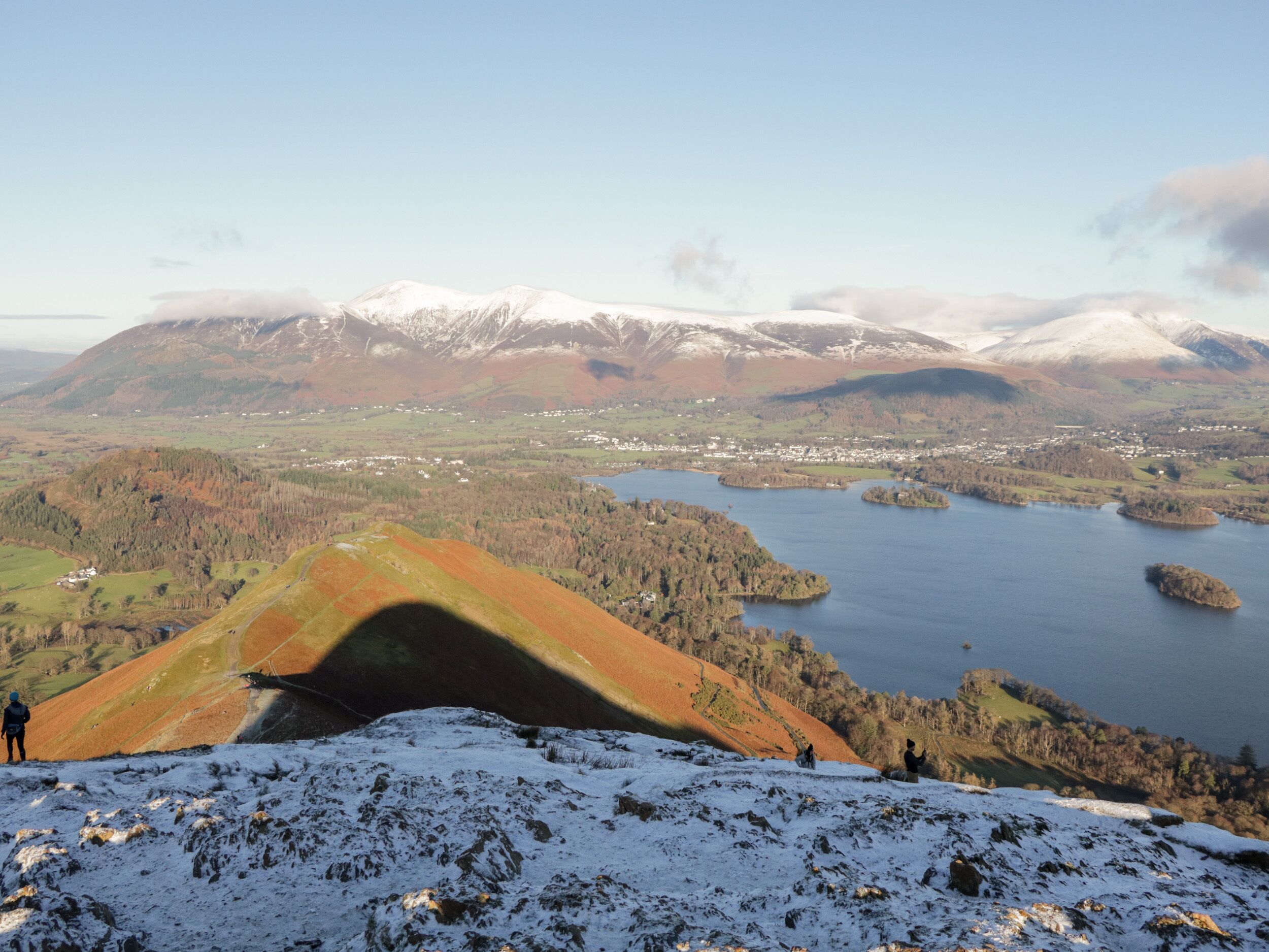 A panoramic view of a mountainous landscape with a lake, snow-capped peaks, and a small settlement in the distance.