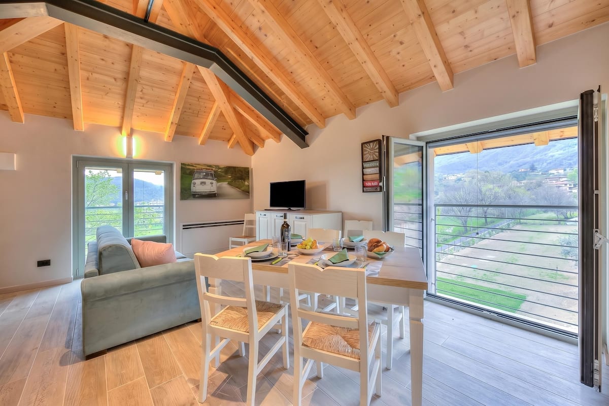 A modern kitchen with a dining area, wooden ceiling beams, a large window, and a view of the outdoors.