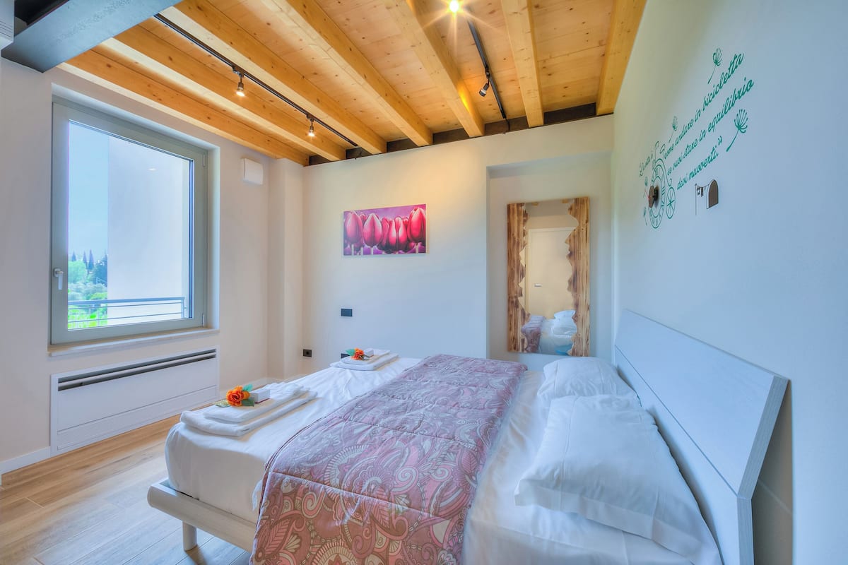 A bedroom with a large window, a bed with a patterned bedspread, a painting on the wall, and a wooden ceiling.