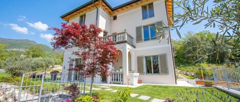 A two-story house with a balcony, a garden with flowers, and a view of the countryside.