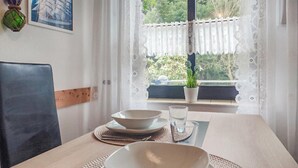 Una mesa de comedor con platos, un vaso y una cuchara, frente a una ventana con cortinas de encaje y vistas al exterior con vegetación.