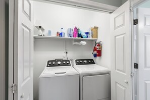 A laundry room with a front-loading washer and dryer, a shelf with cleaning supplies, and a fire extinguisher.