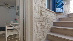 Una escalera con paredes de piedra blanca y contraventanas azules, que conduce a una habitación con una silla blanca y una pequeña mesa con una taza y una flor.