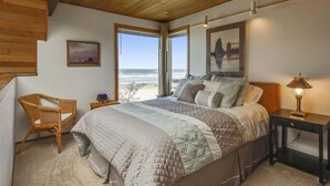 A bedroom with a large bed, a chair, a nightstand, and a view of the beach.