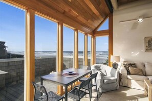 A spacious balcony with a wooden ceiling, large windows offering ocean views, a dining table with chairs, and a sofa. - Ocean House - Oceanfront! (Cannon Beach)