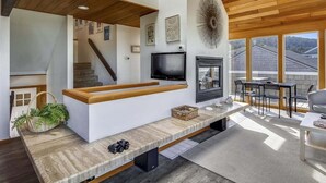 A modern living room with a built-in TV, wooden beams, a fireplace, and a dining area with a view of the outside.