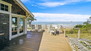 A wooden deck with outdoor seating, a glass door entrance, and a view of the ocean. - Cove Beach Lodge - Magnificent Oceanfront! (Arch Cape)