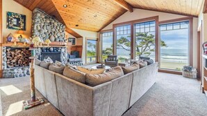 A cozy living room with a stone fireplace, a large sectional sofa, and a view of the ocean. - Cove Beach Lodge - Magnificent Oceanfront! (Arch Cape)