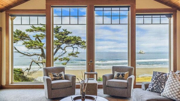 A room with three large windows offering ocean views, two armchairs, a sofa, and a coffee table with a decorative vase. - Cove Beach Lodge - Magnificent Oceanfront! (Arch Cape)