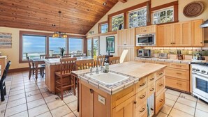A spacious kitchen with wooden cabinets, a large island with a sink, and a view of the ocean through large windows. - Cove Beach Lodge - Magnificent Oceanfront! (Arch Cape)