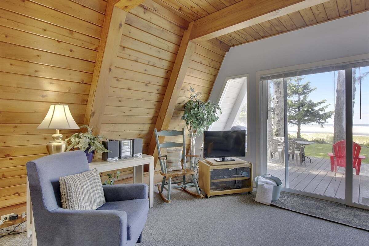 A cozy cabin interior with a wooden ceiling, a sofa, a TV, and a sliding glass door leading to an outdoor deck.