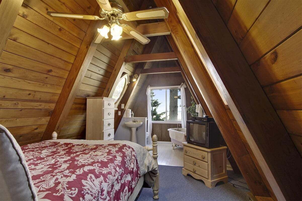 A bedroom with a sloped ceiling, a bed with a floral bedspread, a dresser, a vanity with a mirror, a television, and a window with a view of the outdoors.