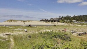Een pand aan het strand met een groot huis en kleinere gebouwen, omgeven door duinen en begroeiing.