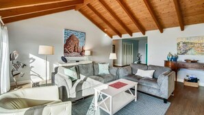 Ein gemütliches Wohnzimmer mit Holzbalkendecke, einer grauen Sofagarnitur, einem weißen Couchtisch und einem Gemälde von einem Berg.