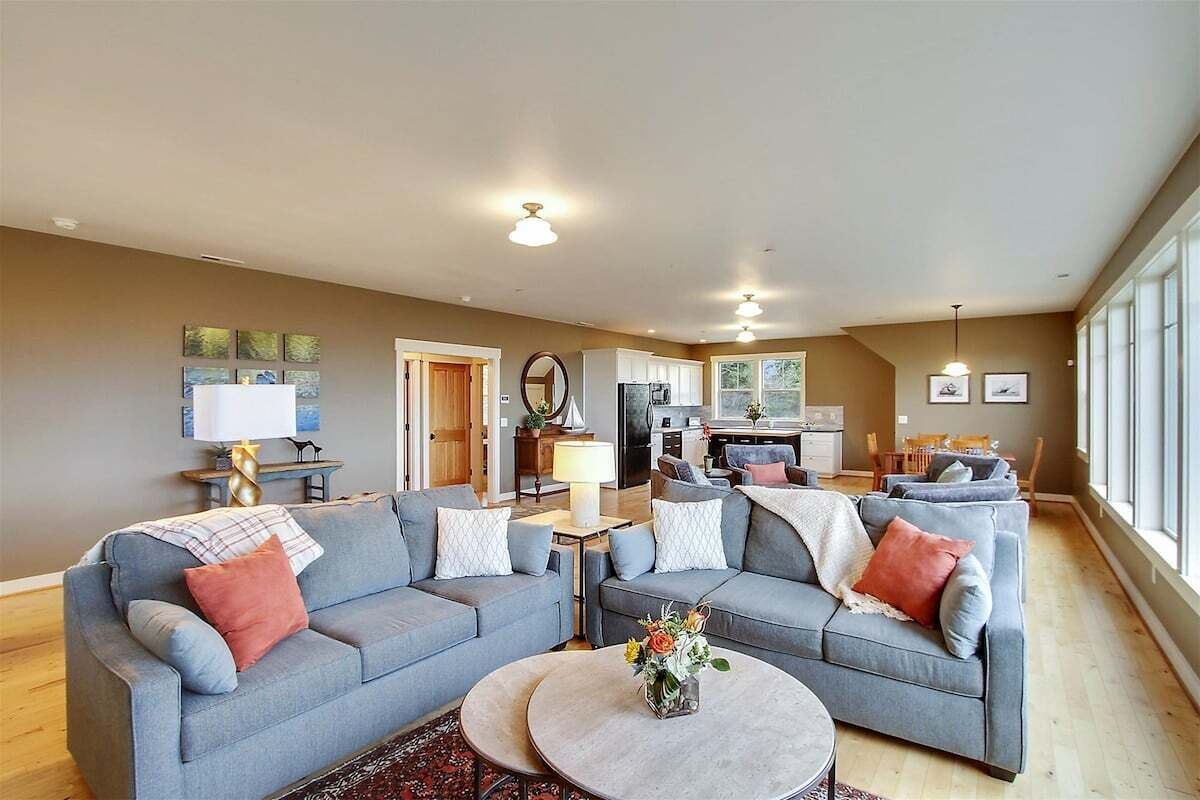 A spacious living room with a grey sofa set, a round coffee table with flowers, and a kitchen in the background.