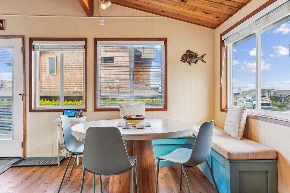 A dining area with a round table, chairs, and a bench with cushions. There are two windows with blinds, a fish decoration on the wall, and a view of the outside.