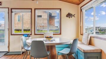 A dining area with a round table, chairs, and a bench with cushions. There are two windows with blinds, a fish decoration on the wall, and a view of the outside.