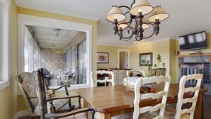 Uma sala de jantar com mesa de madeira e cadeiras estofadas brancas, um candelabro e uma televisão sobre a lareira.
