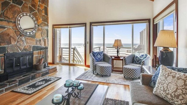 A spacious living room with a stone fireplace, large windows, and a view of the ocean. - Beach Haven - Beachfront! (Cannon Beach)