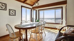 A dining area with a wooden table and chairs, large windows with a view of the ocean, and a hanging light fixture. - Beach Haven - Beachfront! (Cannon Beach)