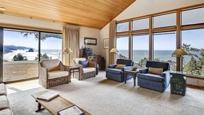 Amplia sala de estar con ventanas grandes que ofrecen vistas al océano. Varias opciones de asiento, incluyendo una silla de mimbre, un sofá azul y un sillón azul. Una mesa de centro de madera está en el centro.