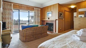 Un cuarto de hotel con una cama grande, un sofá de mimbre con cojines, una chimenea, un televisor y vistas al exterior.