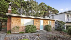 Une maison de plain-pied avec une façade en briques, une cheminée et un petit jardin devant, agrémenté d’un rocail et d’un arbre.