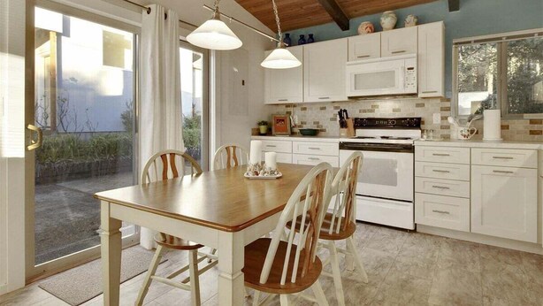 Une cuisine avec des meubles blancs, une table à manger en bois et des chaises. Une fenêtre offre une vue sur la verdure à l’extérieur.