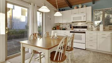 A kitchen with white cabinets, a wooden dining table, and chairs. There is a window with a view of greenery outside.