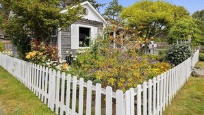 A wooden house with a white picket fence, surrounded by a garden with various plants and flowers.