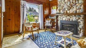 A cozy room with wooden walls, a stone fireplace, a bookshelf, and a view of the outdoors.
