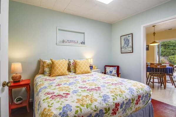 A bedroom with a floral bedspread, a bedside table with a lamp, a framed picture on the wall, and a view of the outdoors through an open door. - Singing Sands - Built in 1920! (Cannon Beach)