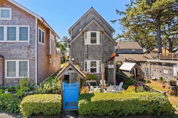 A two-story house with a small front porch, a blue gate, and a garden with various plants and flowers. - Newly Renovated+Fire Table+Steps from Cannon Beach (Cannon Beach)