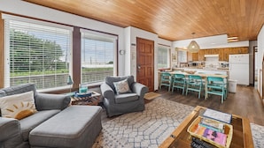 A spacious living area with a sofa, armchair, and coffee table, a kitchen with wooden cabinets, and large windows with blinds. - Shutterbug - 1 Block to the Beach! (Arch Cape)
