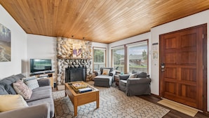 A living room with a stone fireplace, wooden ceiling, grey sofas, a coffee table, and a television. - Shutterbug - 1 Block to the Beach! (Arch Cape)