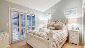 A bedroom with a large bed, two bedside tables, a window with blinds, and a view of the outside.